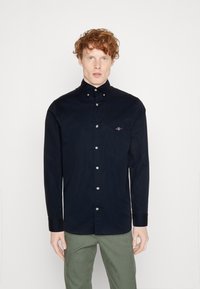 Donkerblauw button-up shirt van gladde stof, met een zak op de linkerkant van de borst en witte knopen aan de voorkant. Lange mouwen.