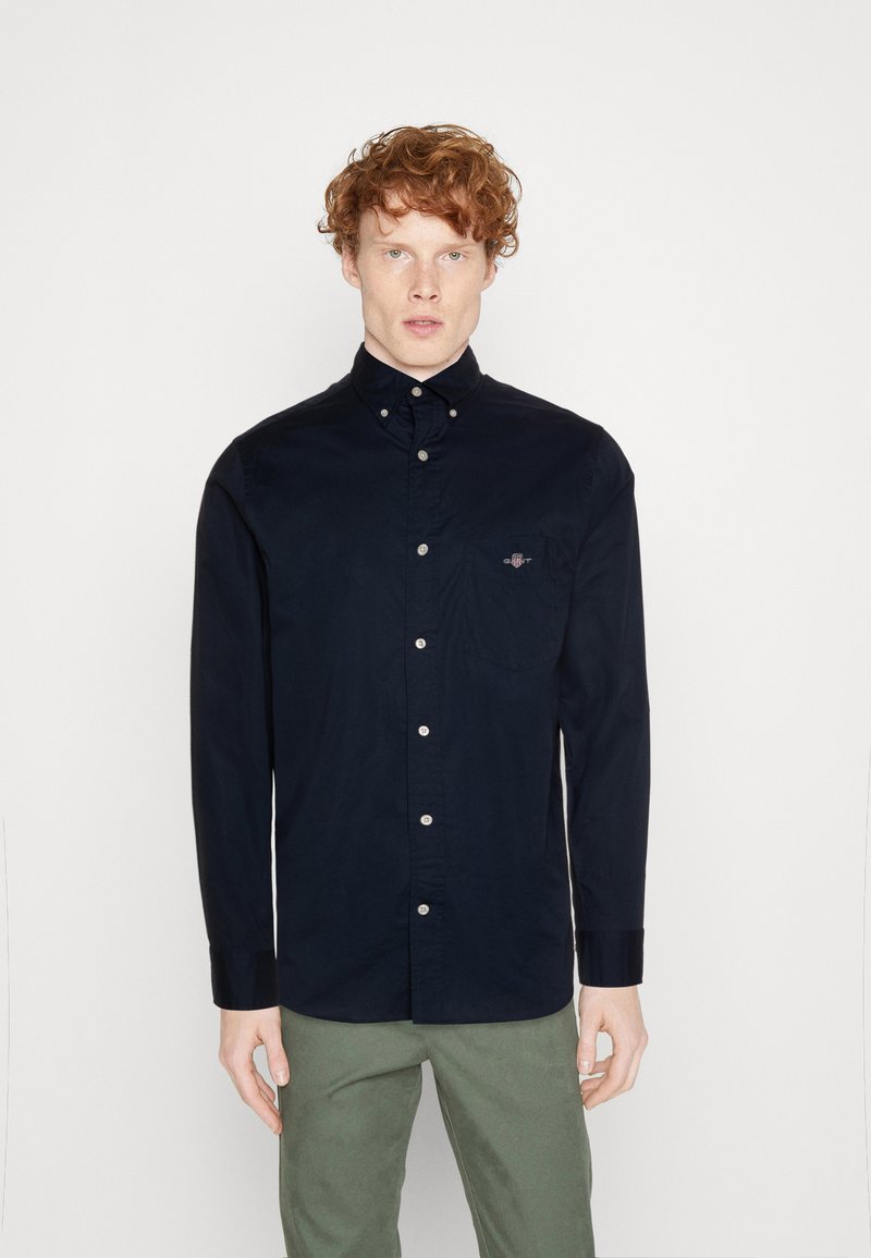 Donkerblauw button-up shirt van gladde stof, met een zak op de linkerkant van de borst en witte knopen aan de voorkant. Lange mouwen.