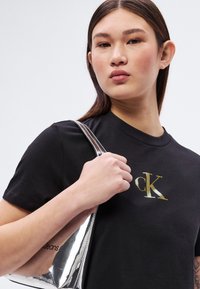 Jeune femme aux cheveux raides portant un t-shirt noir Calvin Klein et tenant un sac à bandoulière argent métallisé sur un fond clair.