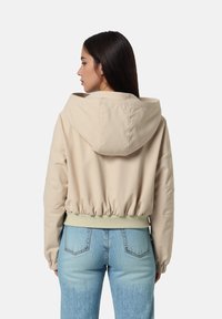 Beige kort jacka med huva, elastiskt midjeband och ribbstickade ärmar, tillverkad av mjukt tyg. I kombination med ljusblå denimjeans.