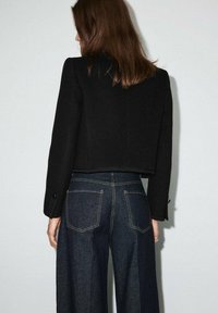 Czarna krótka kurtka z teksturowanej tkaniny, z długimi rękawami i mankietami zapinanymi na guziki, zestawiona z szerokimi jeansami w ciemnym kolorze.