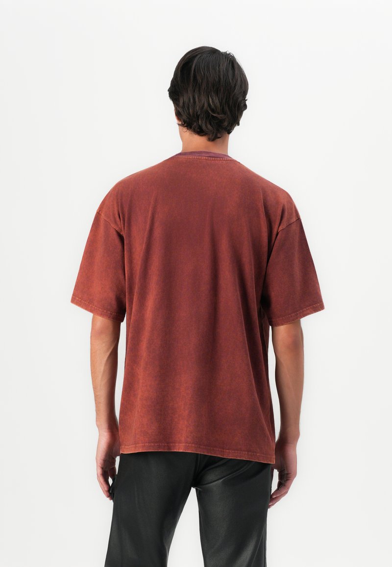 Mężczyzna z ciemnymi włosami, ubrany w luźną, krótką koszulkę w kolorze rdzy i czarne spodnie, stoi przodem do jednolitego białego tła.