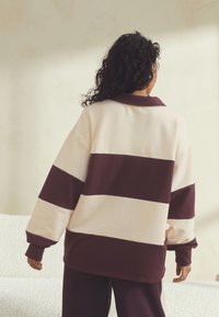 Oversized trui met crème en donker bordeauxrode strepen. Voorzien van een kraag en lange mouwen. Soepele textuur, casual pasvorm, minimalistisch ontwerp.