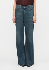 Τζιν παντελόνι σε σκούρο μπλε χρώμα, σχεδίαση με φαρδιά πόδια, με μπροστινές τσέπες και κούμπωμα με κουμπί. Το ύφασμα έχει φθαρμένο υφή με διακριτικές ξεθωριασμένες περιοχές.