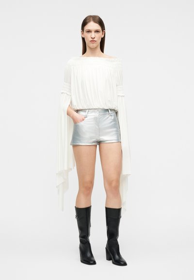 Γυναίκα με λευκό μπλουζάκι χωρίς ώμους με μακριά ρέοντα μανίκια, ασημένια μεταλλικά σορτς και μαύρες μπότες μέχρι το γόνατο, όρθια.