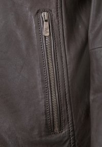 Braune Lederjacke mit einer Reißverschlusstasche. Der Reißverschluss ist silberfarben, und die Textur ist glatt mit sichtbaren Nähten entlang der Kanten.