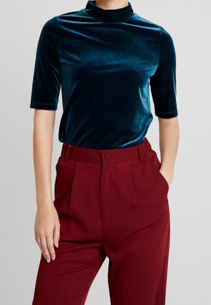 Teal fløjlsbluse med høj mock-hals og korte ærmer, kombineret med højtaljede bordeaux bukser. Blød tekstur og tætsiddende design.