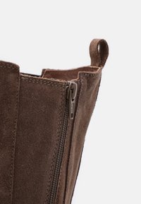 Bottine en daim marron avec une fermeture éclair latérale et un passant. Vue rapprochée du matériau texturé et du détail de la fermeture éclair, mettant en valeur le savoir-faire.