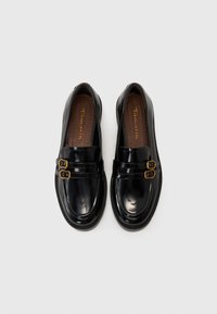 Melnas lakotas ādas loafers ar dubultu sprādzēm. Gluda virsma, noapaļota purngala un mīksts zolītes iešuvums ar perforācijām.