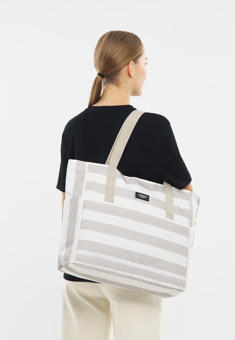 Fekete pólót és krémszínű nadrágot viselő személy egy nagy, bézs és fehér csíkos táskával a vállán.