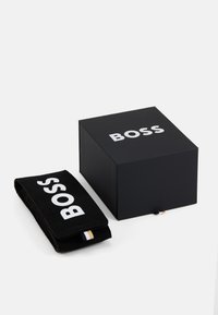 Juodas dežutė su baltu "BOSS" logotipu šalia juodo audinio piniginės su logotipu. Dėžutė pasižymi minimalistiniu dizainu, piniginė turi tekstūruotą paviršių.