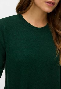 Tmavě zelený svetr s texturovanou tkaninou, kulatým výstřihem a žebrovanými manžetami. Detailní pohled zaměřený na horní část a výstřih.