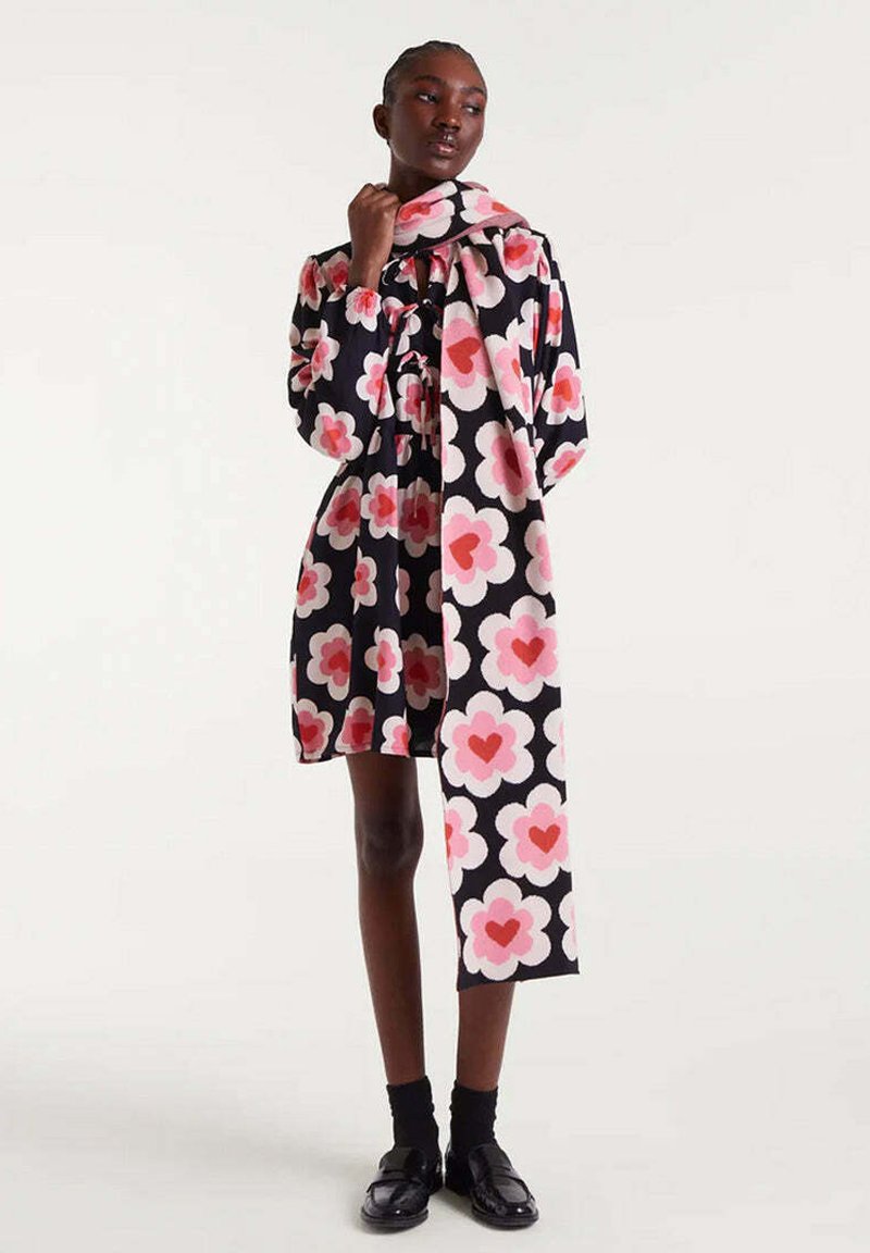Vestido de estampado floral en negro, con grandes flores rosas y blancas con formas de corazón, cintura ceñida y bufanda a juego, combinado con zapatos negros.