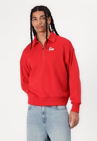 Crvena zip-up dukserica s visokim ovratnikom, koja ima mali bijeli "Gap" logo. Napravljena je od mekane tkanine, a ima rebraste manžete i porub.