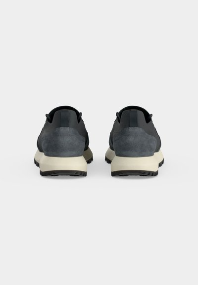 Paire de baskets gris foncé et noir avec des semelles beige, vues de dos sur un fond clair uni.