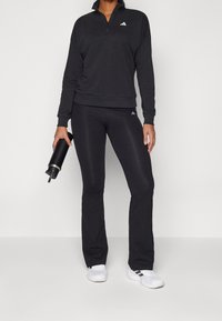 Osoba w czarnej sportowej bluzie na zamek i legginsach trzyma czarną butelkę na wodę, mając na sobie białe trampki z czarnymi paskami.