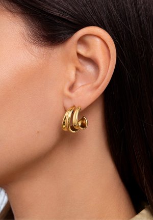 Pendientes en tono dorado que presentan tres formas curvas y superpuestas con un acabado brillante, colocados sobre el lóbulo de la oreja contra un fondo de piel suave.