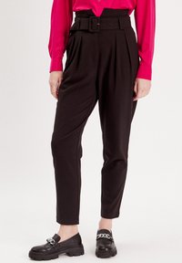 Schwarze hoch taillierte Hosen mit einem Gürtel, gefalteter Vorderseite, schmal zulaufenden Beinen und einer glatten Textur. Tragen mit klobigen schwarzen Slippern.