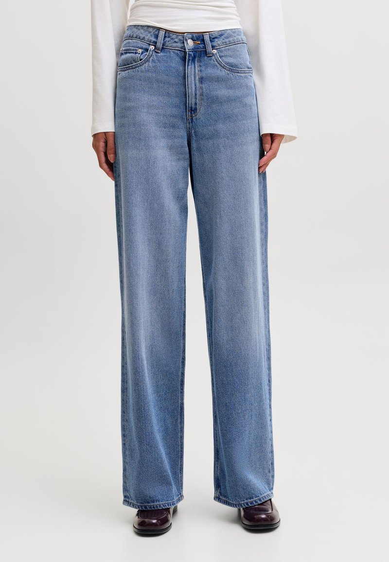 Personne portant un jean large bleu clair, un haut à manches longues blanc et des chaussures foncées, debout devant un fond clair uni.