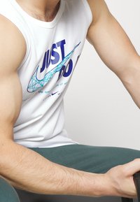 Spierkrachtige persoon in een wit mouwloos Nike-shirt met de tekst "JUST DO IT" en een geometrisch swoosh-ontwerp, die een voorwerp met beide handen vasthoudt.