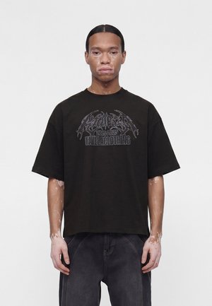 Nuori henkilö, jolla on vitiligo, pukeutuneena mustaan ylisuurikokoiseen graafiseen t-paitaan ja tummiin farkkuihin, seisoo yksinkertaisen vaalean taustan edessä.