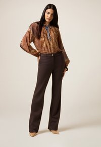 Braune Bluse mit Paisleymuster und blauer Schleife, ergänzt durch dunkelbraune gerade Hosen und beige hochhackige Schuhe.