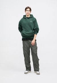 Sweatshirt verde com um bolso canguru e logótipo, emparelhado com calças cargo cinzas com múltiplos bolsos. Ténis com uma combinação de cores branca e cinza.