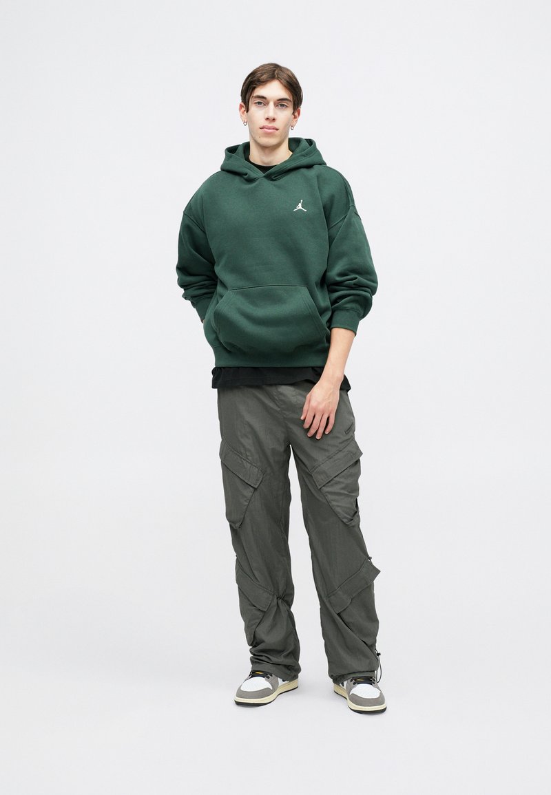 Sweatshirt verde com um bolso canguru e logótipo, emparelhado com calças cargo cinzas com múltiplos bolsos. Ténis com uma combinação de cores branca e cinza.