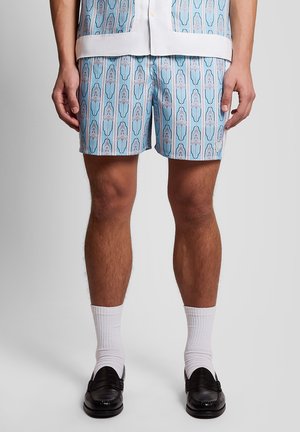 Homme portant un short bleu à motifs, des chaussettes blanches, des mocassins noirs et une chemise assortie avec un ourlet blanc.