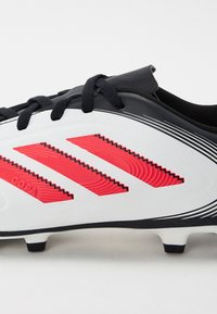 Botas de fútbol blancas con acentos negros, que presentan tres franjas en zigzag rojas. Textura suave, cordones negros y tacos en la suela para tracción.
