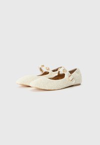 Chaussures plates blanches texturées avec un nez pointu, dotées de deux sangles à boucle et d'accents dorés décoratifs sur la sangle.