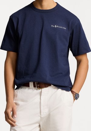 Mann trägt ein marineblaues Polo Ralph Lauren T-Shirt, beige Hose, braunen Ledergürtel und eine Armbanduhr mit dunklem Zifferblatt am linken Handgelenk.
