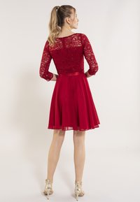 Robe rouge avec un corsage en dentelle, des manches trois-quarts et une jupe évasée. Finition avec une ceinture en satin et portée avec des talons métalliques.