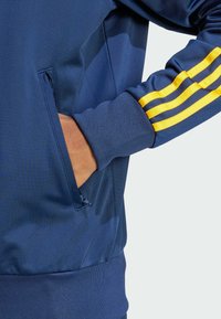 Navy plavi dukserica s patentnim zatvaračem na džepu, rebrastim manžetama i tri žute horizontalne pruge na rukavu. Glatka tekstura.