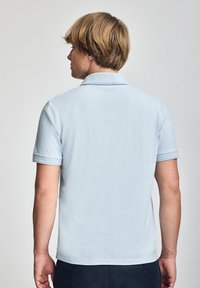 Polo de color azul claro con una textura suave, mangas cortas y un cuello clásico; costuras visibles en las mangas y el dobladillo.