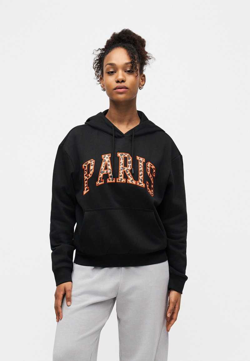 Νεαρή γυναίκα με σγουρά μαλλιά, φορώντας μαύρο φούτερ με κουκούλα με την επιγραφή "PARIS" και ανοιχτό γκρι παντελόνι, στέκεται μπροστά σε απλό λευκό φόντο.