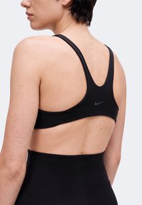 Άτομο που φοράει μαύρο αθλητικό σουτιέν Nike με σχέδιο racerback, εμφανίζεται από πίσω σε απλό ανοιχτό φόντο.