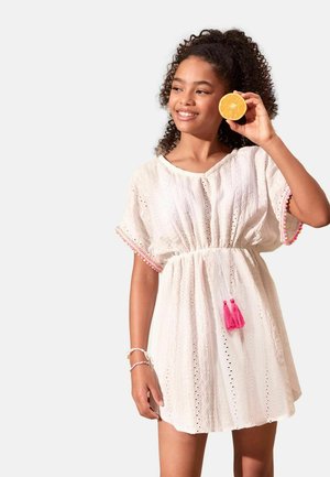 Mädchen mit lockigem Haar im weißen Kleid, hält eine halbe Orange neben ihren Kopf, lächelt und schaut seitlich vor neutralem Hintergrund.