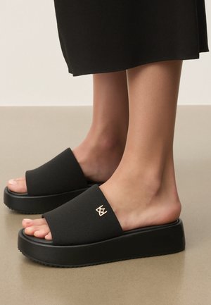 Fødder iført sorte platform-slide-sandaler med bred tekstureret rem og gulddetalje med logo på en beige gulv.