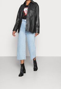 Persona che indossa una giacca in pelle nera con bottoni, una maglietta nera con logo, jeans a gamba larga blu chiaro tagliati alla caviglia e stivali neri con tacco.