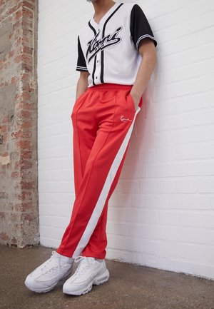 Person lehnt an einer weißen Ziegelwand und trägt ein schwarz-weißes Baseballshirt, rote Jogginghosen mit weißen Streifen und weiße Turnschuhe.