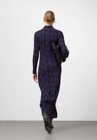 Langes karierte Kleid in Dunkelblau und Burgunderrot mit enger Silhouette und langen Ärmeln, getragen mit einer schwarzen Tasche und eleganten, heeled Stiefeletten.