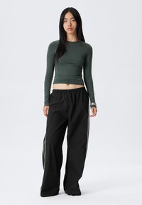 Long-sleeve dark green fitted top paired with loose black pants featuring white side stripes. Smooth fabric with a crew neckline.