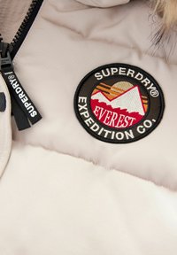 Beige Steppjacke mit einem runden gestickten Patch, das Berge und den Text "SUPERDRY EXPEDITION CO. EVEREST" zeigt. Schwarze Reißverschlussdetails.