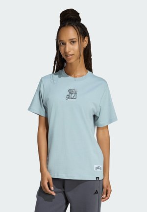 Donna con i capelli intrecciati che indossa una maglietta azzurro chiaro con una piccola grafica nera e pantaloni Adidas grigio scuro, in piedi contro uno sfondo semplice.