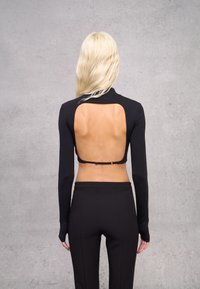 Top nero a maniche lunghe con grande apertura sulla schiena, design aderente e collo alto. La cucitura accurata e la trama liscia sono visibili.