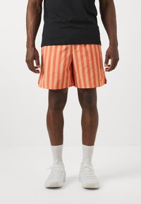 Bañador a rayas en naranja y coral, de longitud hasta la rodilla, fabricado en tejido ligero, con aberturas laterales, combinado con zapatillas y calcetines blancos.