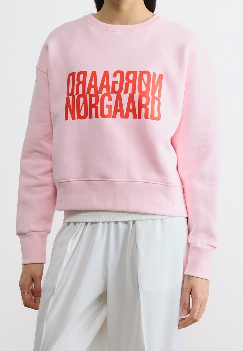 Personne portant un sweat-shirt rose avec un texte rouge "NØRGAARD" et un pantalon blanc ample, debout devant un fond uni.