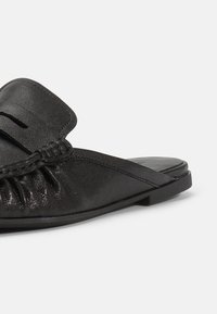 Μαύρο δερμάτινο loafers χωρίς πίσω μέρος με πλεκτή λεπτομέρεια και ελαφρές ρυτίδες στο μπροστινό μέρος, με επίπεδη σόλα, πλευρική όψη σε λευκό φόντο.
