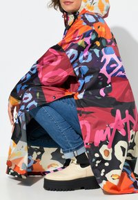 Poncho imperméable coloré avec un motif léopard et abstrait, doté d'une capuche, de boutons-pression et d'une coupe ample sur des jeans et des bottes.